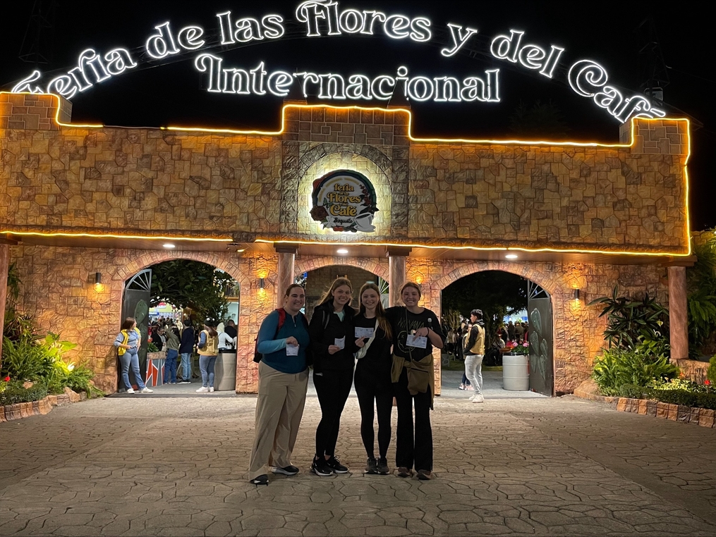 Four people standing in front of the illuminated entrance to the Feria de las Flores y del Café festival at night.